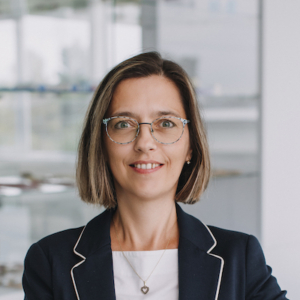 Portrait of a smiling professional woman with short brown hair and glasses, wearing a navy blazer with white piping in a bright office setting.